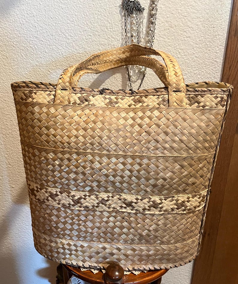 K&ouml;nnte beinhalten: Eine gro&szlig;e, gewebte Tragetasche aus nat&uuml;rlichen Materialien. Die Tasche hat eine rechteckige Form mit einer breiten Basis und einem schmalen oberen Teil. Die Tasche ist aus einem hellbraunen Material mit einem gewebten Muster gefertigt. Die Tasche hat zwei Henkel, die in die Tasche eingewebt sind.