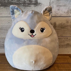 Puede incluir: Un juguete de peluche azul claro con forma de zorro con detalles blancos. El juguete tiene una cara sonriente con ojos negros y una lengua rosa.