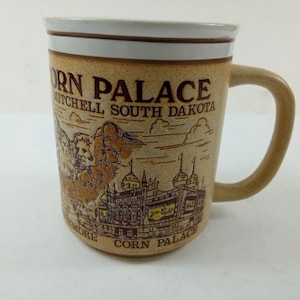 Könnte beinhalten: Beige Keramikbecher mit braunem Henkel und Rand. Der Becher trägt die Aufschrift "Corn Palace Mitchell South Dakota" und eine Illustration des Corn Palace Gebäudes und des Mount Rushmore.