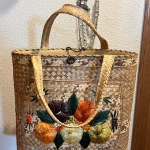 K&ouml;nnte beinhalten: Eine geflochtene Strohtasche mit Blumenmuster. Die Blumen sind mit buntem Garn gestickt und in den Farben Orange, Gelb, Gr&uuml;n und Braun gehalten.