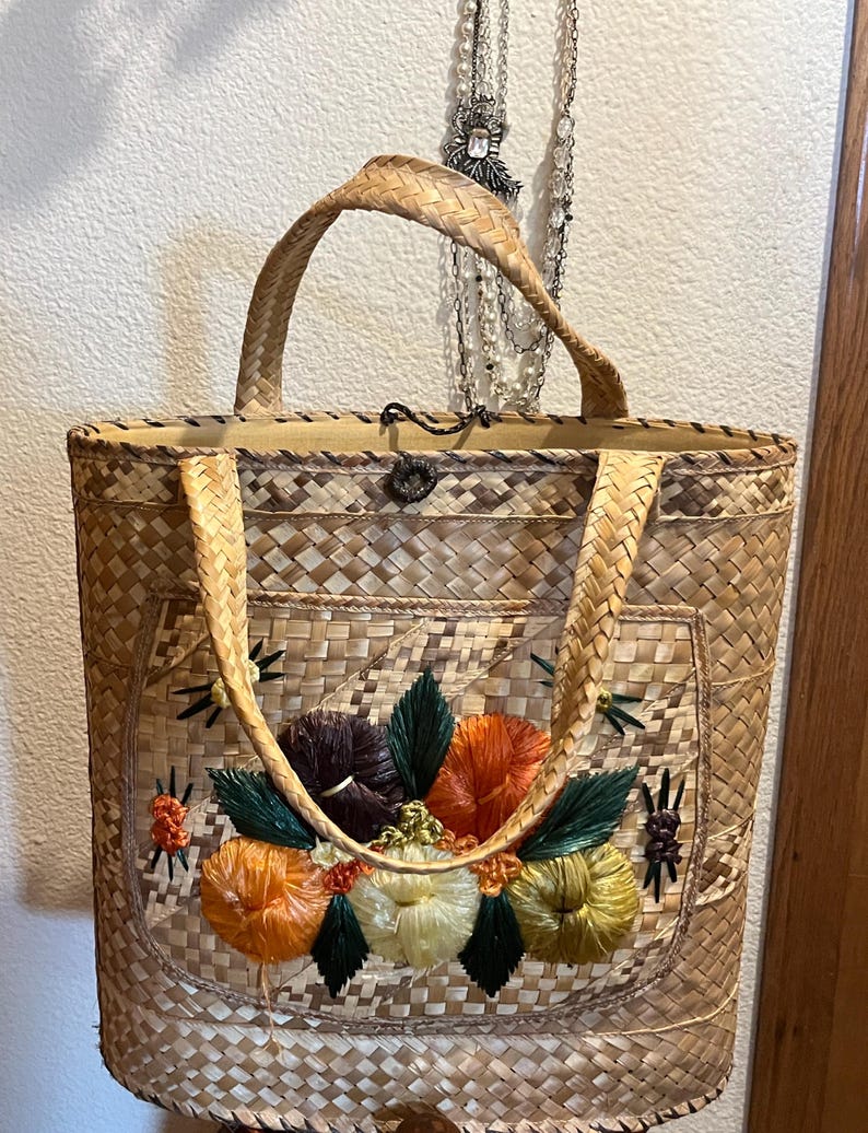 K&ouml;nnte beinhalten: Eine geflochtene Strohtasche mit Blumenmuster. Die Blumen sind mit buntem Garn gestickt und in den Farben Orange, Gelb, Gr&uuml;n und Braun gehalten.