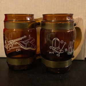 May include: Two brown glass mugs with gold metal bands around the top and bottom. The mugs have a wooden handle and are decorated with white images of cowboy boots, a gun, and a holster. The text "M-V", "DR", "K", and "B" are printed on the mugs.