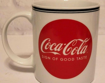 Op de afbeelding: Witte keramische mok met een rode cirkel en het Coca-Cola-logo in witte letters. De tekst "Sign of Good Taste" staat onder het logo.