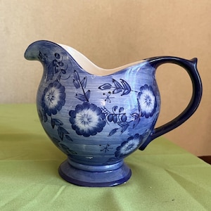Op de afbeelding: Een blauwe en witte keramische roomkan met een bloemenpatroon. De roomkan heeft een handvat en een tuit.