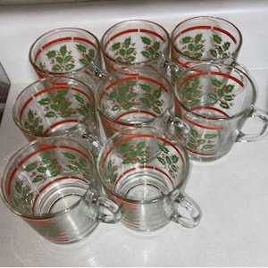 May include: Set of eight clear glass mugs with red and green holly and berry designs. The mugs have a red band around the top and a handle.