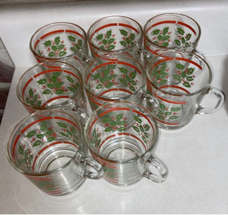 May include: Set of eight clear glass mugs with red and green holly and berry designs. The mugs have a red band around the top and a handle.