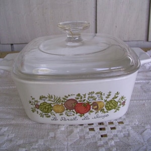 Puede incluir: Plato de cerámica blanco para hornear con tapa de vidrio. El plato tiene un diseño floral verde, marrón y rojo con el texto "Le Persil La Sauge".