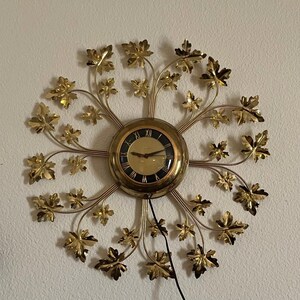 May include: A gold-toned wall clock with a circular face and a decorative leaf design. The clock face has black numerals and hands. The clock is surrounded by gold-colored metal leaves and stems, creating a sunburst effect. A black power cord is visible.