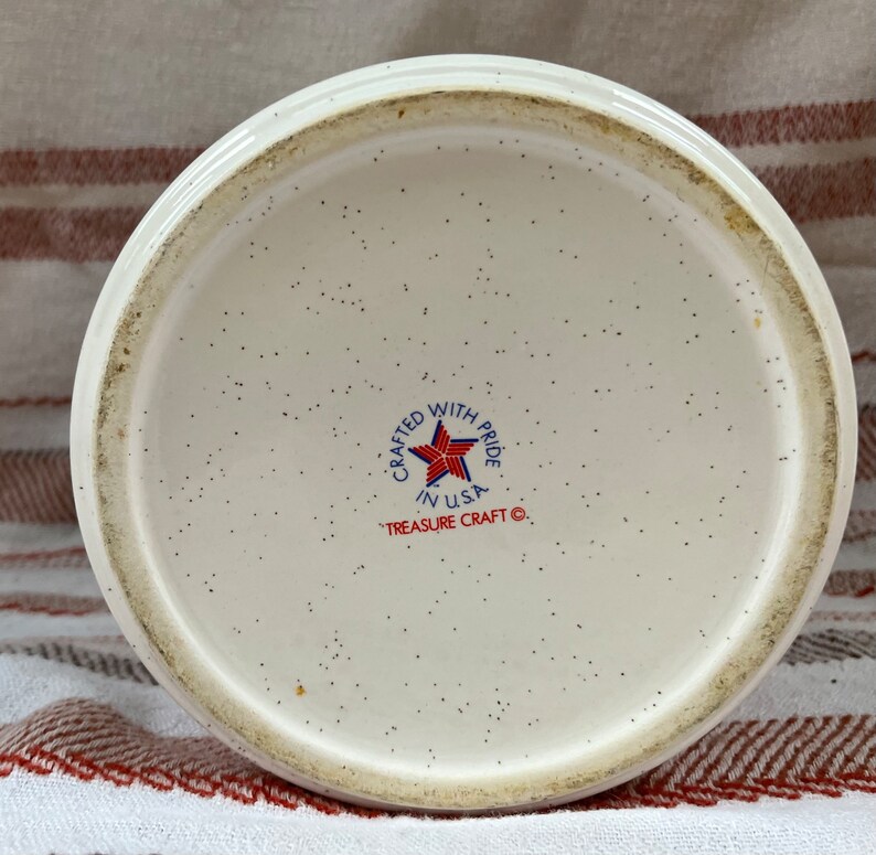 Puede incluir: La parte inferior de una taza de cer&aacute;mica blanca con un dise&ntilde;o de estrella roja, blanca y azul. El texto "Crafted with Pride in USA" y "Treasure Craft &copy;" est&aacute; impreso en la taza.