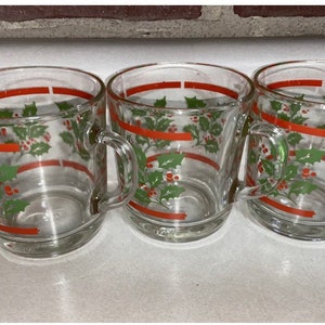 May include: Three clear glass mugs with red and green holly leaf and berry designs. The mugs have handles.