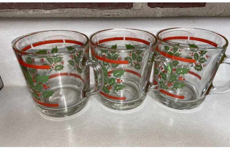 May include: Three clear glass mugs with red and green holly leaf and berry designs. The mugs have handles.