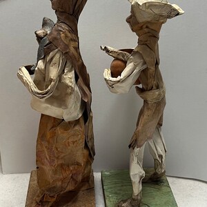 May include: Two vintage paper mache figurines of a man and woman, both wearing traditional clothing. The man is holding a brown pot and the woman is holding a small animal. They are both standing on wooden bases.