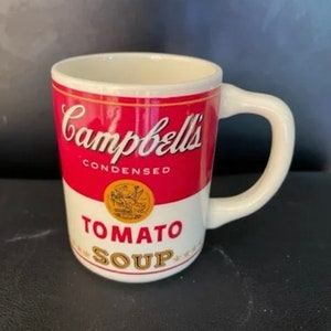 Könnte beinhalten: Eine weiße Keramiktasse mit einem roten und gelben Design einer Campbell's Tomato Soup-Dose. Die Tasse hat einen weißen Henkel und der Text "Campbell's Condensed Tomato Soup" ist auf der Vorderseite aufgedruckt.