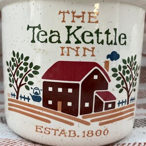 Puede incluir: Taza de cer&aacute;mica blanca con una ilustraci&oacute;n marr&oacute;n y roja de una casa con un techo rojo y una chimenea. El texto "The Tea Kettle Inn Estab. 1806" est&aacute; impreso en la taza.