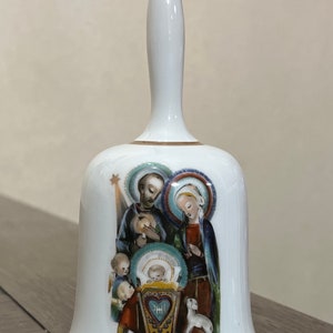 Può includere: Campana in porcellana bianca con bordo dorato e una rappresentazione della Sacra Famiglia. La campana presenta uno sfondo blu e verde con una stella e un agnello. Il testo "Hummel" è visibile in basso.