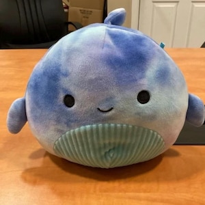 May include: A blue and purple tie-dye plush whale with a light blue belly and a smiling face. The whale has a small fin on top of its head.