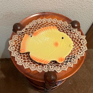 May include: A yellow ceramic fish dish with orange accents sits on a wooden tiered stand with a crocheted doily.