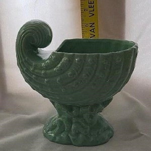 May include: A green ceramic planter shaped like a seashell with a curled lip and a detailed base. The planter is designed to resemble a conch shell.