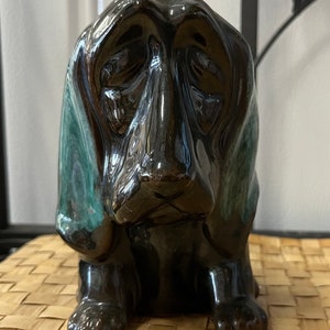 Puede incluir: Una figurita de cerámica de un perro basset negro y verde sentado con la cabeza gacha. El perro tiene un acabado brillante.