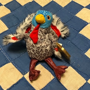 Puede incluir: Un juguete de peluche de pavo con cabeza azul, papada roja y cuerpo de estampado de leopardo gris y marrón. El pavo tiene una cola de plumas blancas y negras y está sentado sobre una tela a cuadros azules y blancos.