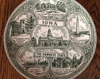 May include: A white ceramic plate with a green border and a design featuring landmarks of Iowa. The plate features the text "IOWA" and "THE HAWKEYE STATE".