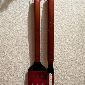 Könnte beinhalten: Zwei Grillwerkzeuge mit Holzgriffen, die an einer weißen Wand hängen. Die Werkzeuge sind ein Spatel und eine Gabel. Der Spatel hat eine rote Metallklinge mit Löchern. Die Gabel hat eine silberne Metallklinge.