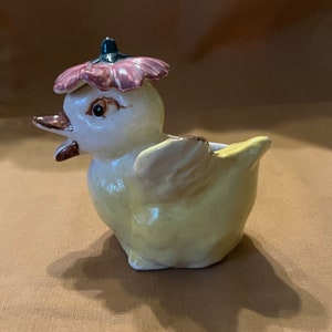 May include: A yellow ceramic chick figurine with a pink flower hat. The chick has a brown beak and black eyes. The figurine is a vintage style.