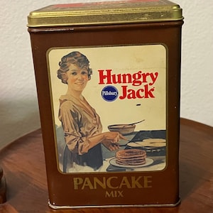 May include: A vintage brown tin box with a gold lid. The front of the box features a woman in a brown dress holding a spatula and a bowl. The text on the box reads "Hungry Jack Pancake Mix" with the Pillsbury logo.