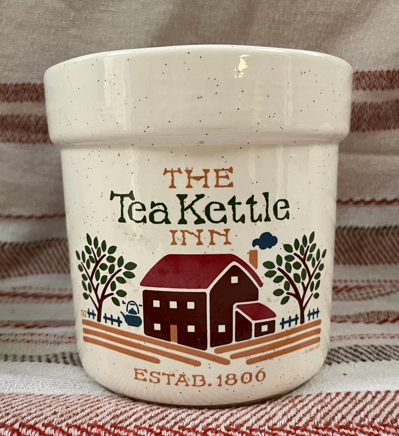 Puede incluir: Jarr&oacute;n de cer&aacute;mica blanco con una ilustraci&oacute;n marr&oacute;n y roja de una casa con techo rojo y chimenea. El texto "THE Tea Kettle INN ESTAB. 1800" est&aacute; impreso en el jarr&oacute;n.