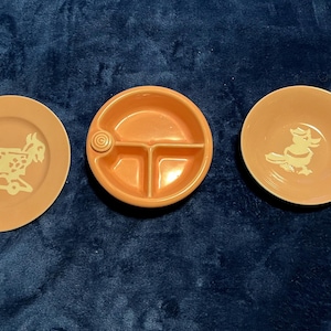 Pode incluir: Três pratos infantis vintage cor de pêssego. Um prato apresenta uma cabra num cavalo de baloiço, outro um pato, e o terceiro é uma tigela dividida. Os pratos são feitos de cerâmica.