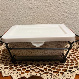 Könnte beinhalten: Eine weiße Keramik-Backform mit Blumenmuster und einem weißen Deckel, die auf einem schwarzen Metallgestell steht. Die Form steht auf einer gehäkelten Unterlage.