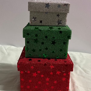 Könnte beinhalten: Drei übereinander gestapelte Geschenkboxen mit roter, grüner und silberner Glitzer-Oberfläche. Jede Box ist mit einem Sternenmuster verziert.