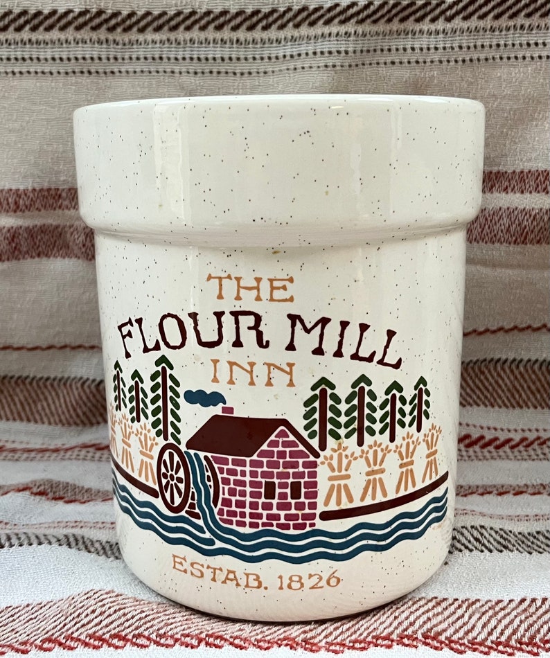 Puede incluir: Jarr&oacute;n de cer&aacute;mica blanca con una ilustraci&oacute;n marr&oacute;n, roja y verde de un molino de harina con el texto "The Flour Mill Inn Estab. 1826".