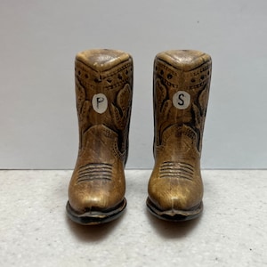 May include: A pair of brown ceramic cowboy boot salt and pepper shakers. The boots are detailed with stitching and have a brown leather look. The shakers are labeled "P" and "S" for pepper and salt.