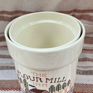 Puede incluir: Bote de cer&aacute;mica blanca con acabado moteado. El bote tiene tapa y presenta una ilustraci&oacute;n vintage de un molino con el texto "The Flour Mill Inn".