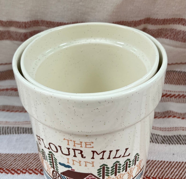 Puede incluir: Bote de cer&aacute;mica blanca con acabado moteado. El bote tiene tapa y presenta una ilustraci&oacute;n vintage de un molino con el texto "The Flour Mill Inn".