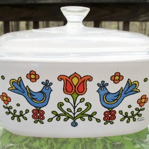 Puede incluir: Plato para hornear blanco con tapa de vidrio que presenta un diseño floral con pájaros azules, tulipanes rojos y hojas verdes. El plato tiene un fondo blanco con un patrón floral colorido.