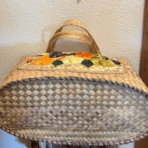K&ouml;nnte beinhalten: Eine geflochtene Strohtasche mit Blumenmuster. Die Tasche ist aus nat&uuml;rlichen Materialien gefertigt und hat einen einzigartigen Vintage-Stil.