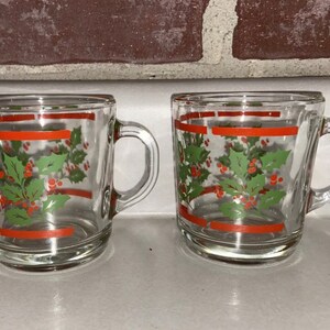 May include: Two clear glass mugs with red and green holly designs. The mugs have a red stripe around the top and bottom.