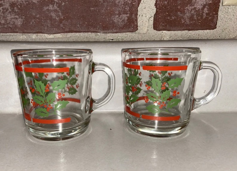 May include: Two clear glass mugs with red and green holly designs. The mugs have a red stripe around the top and bottom.