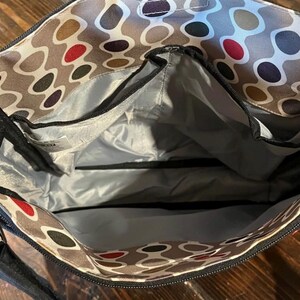 Puede incluir: Vista abierta de un bolso tote estampado con m&uacute;ltiples compartimentos. El interior del bolso es gris, con cremalleras y ribetes negros. El exterior presenta un patr&oacute;n repetitivo de &oacute;valos marrones, rojos y negros sobre un fondo gris claro. El bolso tiene una correa negra para el hombro.