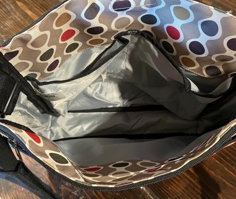 Puede incluir: Vista abierta de un bolso tote estampado con m&uacute;ltiples compartimentos. El interior del bolso es gris, con cremalleras y ribetes negros. El exterior presenta un patr&oacute;n repetitivo de &oacute;valos marrones, rojos y negros sobre un fondo gris claro. El bolso tiene una correa negra para el hombro.