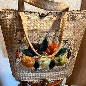 K&ouml;nnte beinhalten: Eine geflochtene Strohtasche mit einem gestickten Blumendesign auf der Vorderseite. Die Tasche hat zwei lange Henkel und einen Klappverschluss.