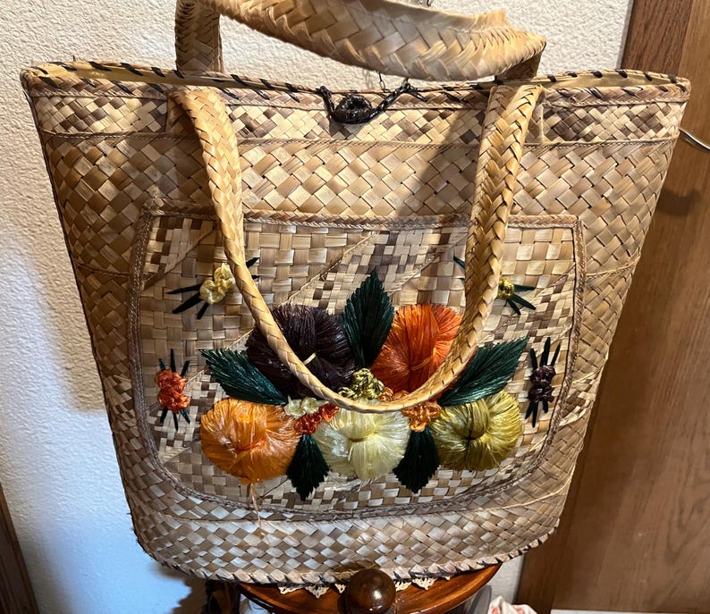 K&ouml;nnte beinhalten: Eine geflochtene Strohtasche mit einem gestickten Blumendesign auf der Vorderseite. Die Tasche hat zwei lange Henkel und einen Klappverschluss.