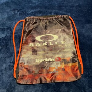 May include: A drawstring bag with the Oakley logo in white and the text "Buckle B" in red. The bag has a cityscape design in shades of orange, brown, and black, with orange drawstrings.