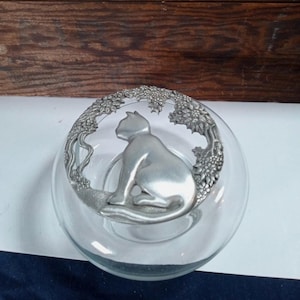 Puede incluir: Un cuenco de vidrio transparente con una figura de gato plateada en su interior. El gato está rodeado por un marco metálico decorativo con un diseño floral. El cuenco es redondo y se apoya sobre una superficie blanca y azul.