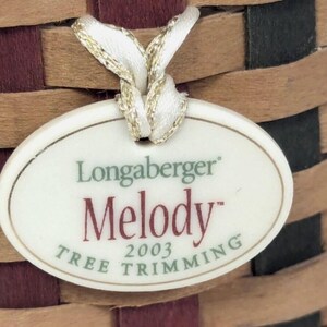 Könnte beinhalten: Nahaufnahme eines geflochtenen Korbes mit einem Etikett. Der Korb besteht aus hellbraunen und dunkelbraunen gewebten Streifen mit burgunderroten und schwarzen Akzenten. Auf dem Etikett steht "Longaberger Melody Tree Trimming 2003".