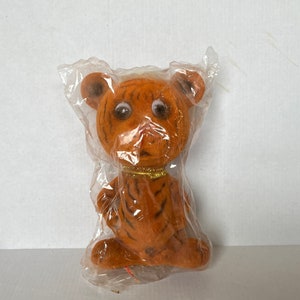 Op de afbeelding: Een pluche tijger speelgoed met oranje en zwarte strepen, witte ogen en een gouden kraag. Het speelgoed is verpakt in doorzichtig plastic.