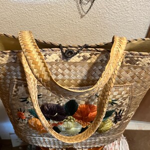 K&ouml;nnte beinhalten: Eine geflochtene Strohtasche mit gestickten Blumenmotiven in Orange-, Gr&uuml;n- und Lilat&ouml;nen. Die Tasche hat zwei geflochtene Henkel und einen schwarzen Kordelverschluss.