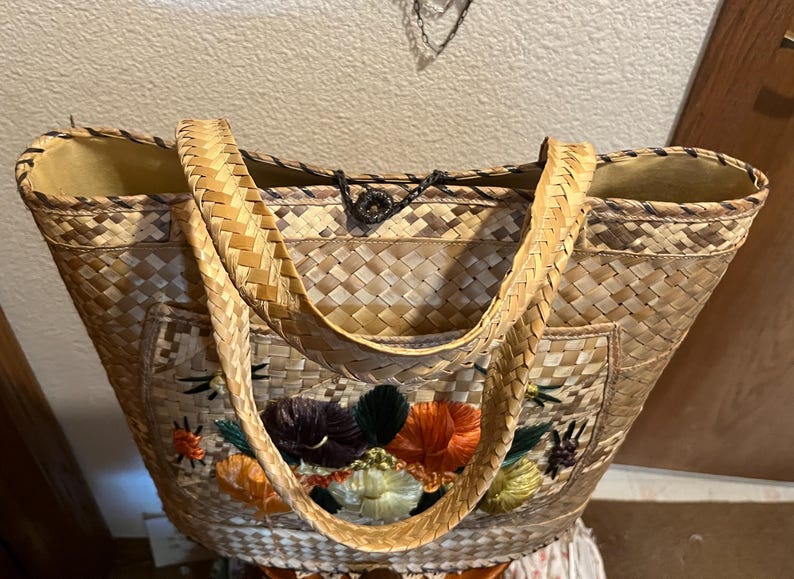 K&ouml;nnte beinhalten: Eine geflochtene Strohtasche mit gestickten Blumenmotiven in Orange-, Gr&uuml;n- und Lilat&ouml;nen. Die Tasche hat zwei geflochtene Henkel und einen schwarzen Kordelverschluss.